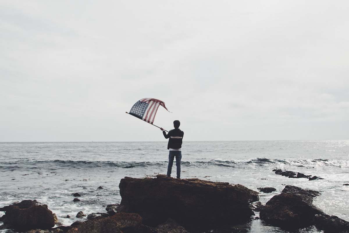 grey man holding flag of America standing on boulder near seashore water